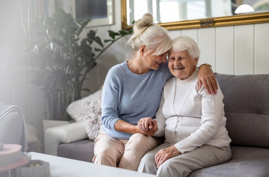 Middle-aged woman with white hair hugging and holding hand of older woman in white cardigan on a couch. Both are smiling.