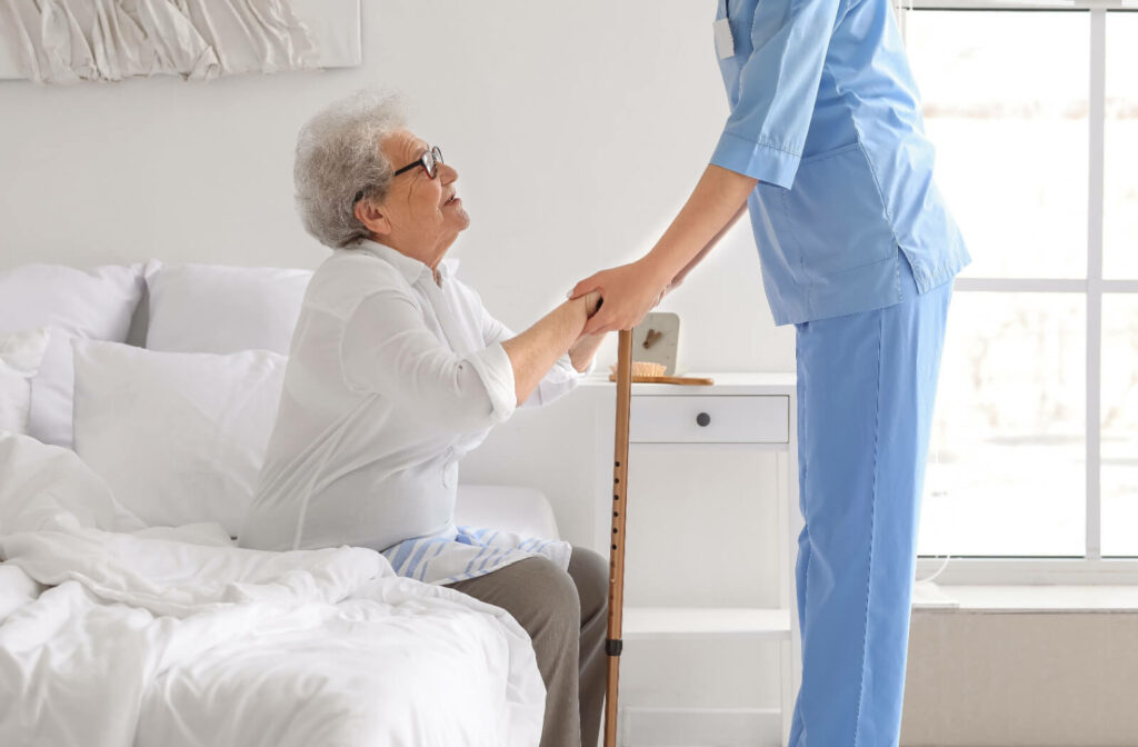 An out-of-frame caregiver helps an older resident with a cane prepare to stand up from their bed in senior living