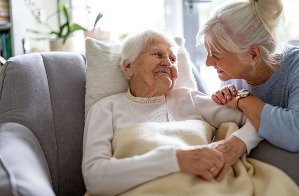 A senior smiling while laying comfortably on a sofa chair with a caregiver resting on the arm